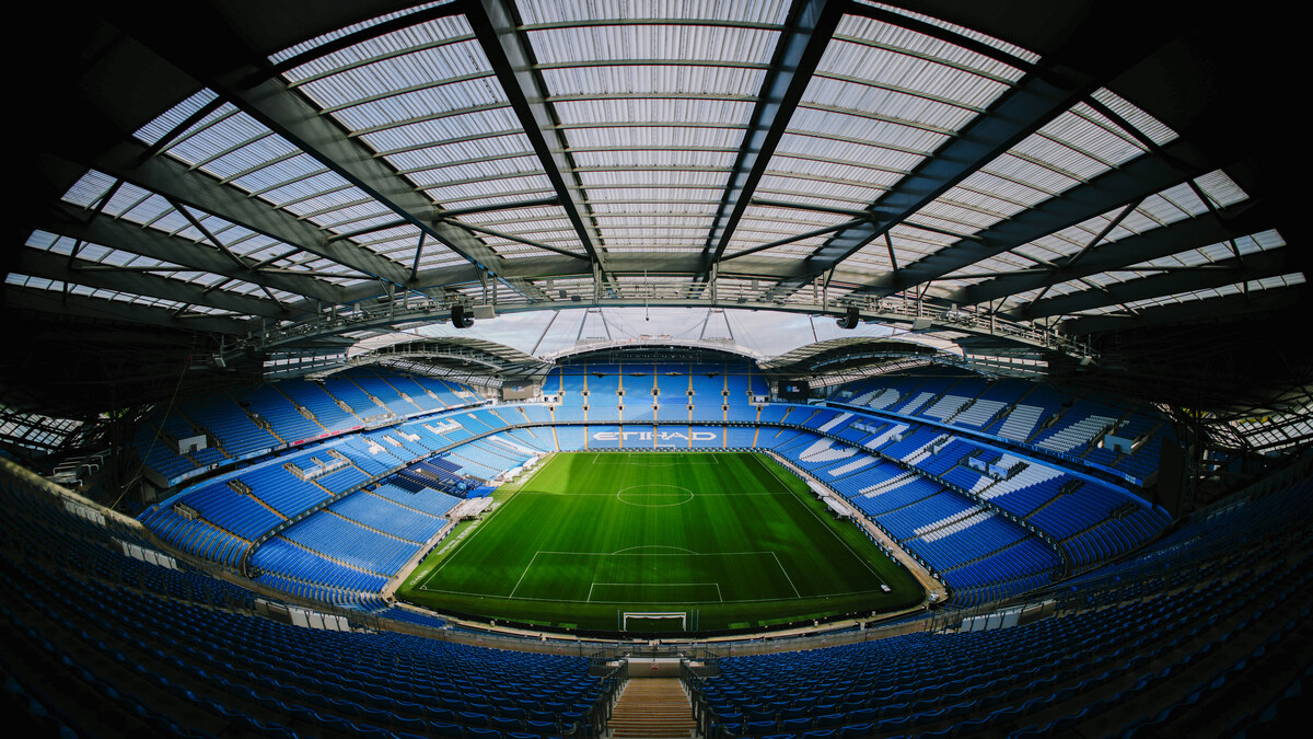 A wide interior view of Manchester City's Etihad Stadium with the pitch and blue seating fully visible
