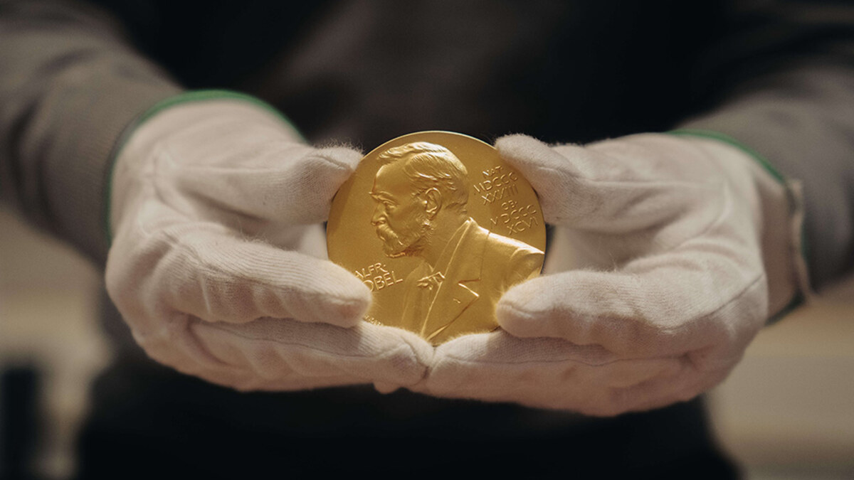 A Nobel Prize medal displayed beside the official blue-and-gold Nobel announcement backdrop